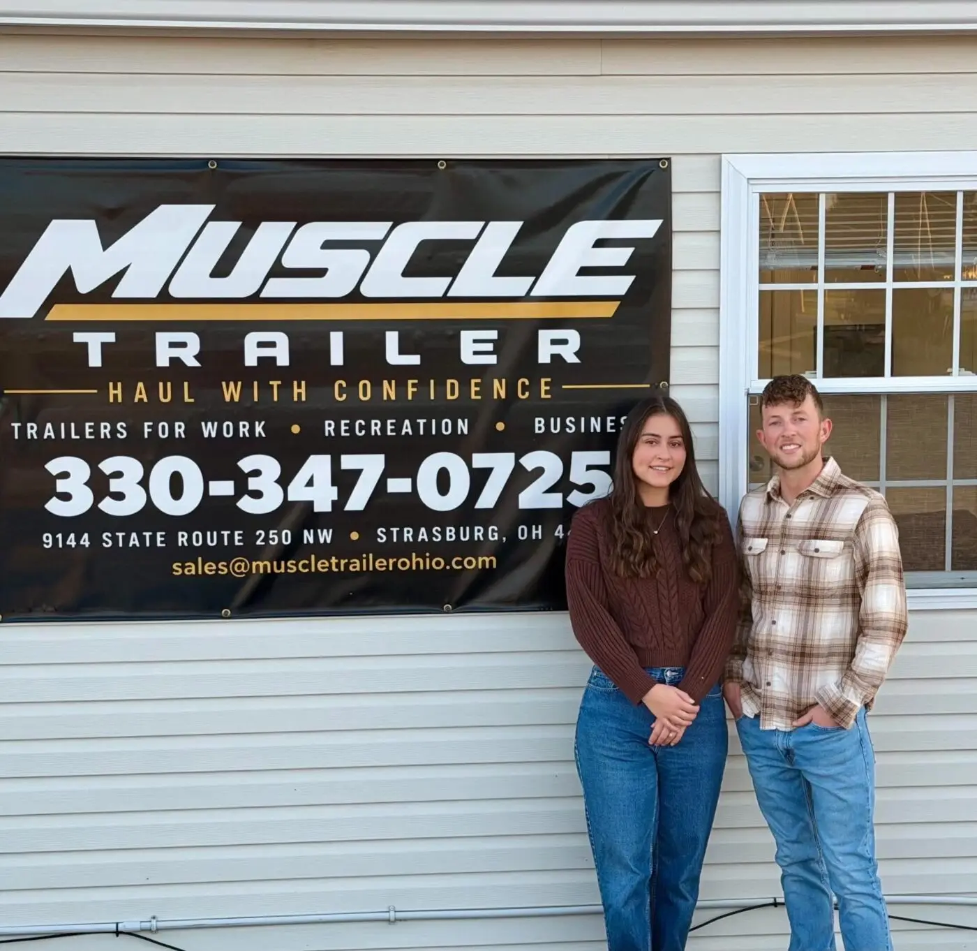 Two people stand beside a Muscle Trailer business banner.